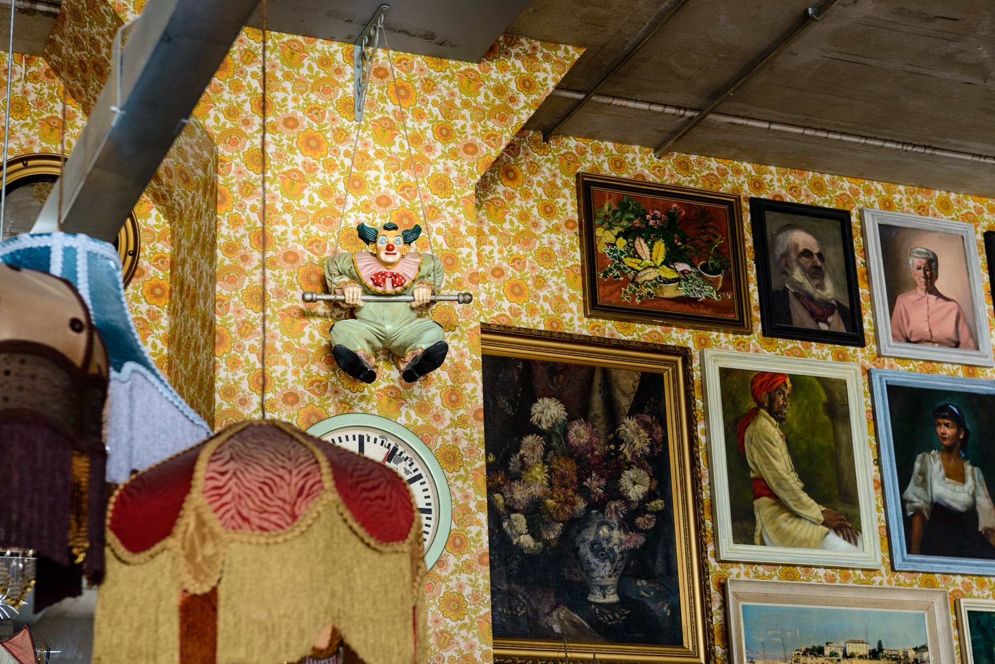 A vintage room with floral wallpaper, framed paintings, and a Visto clock on the wall. A clown doll sits on a trapeze, while a fringed lampshade hangs in the foreground.