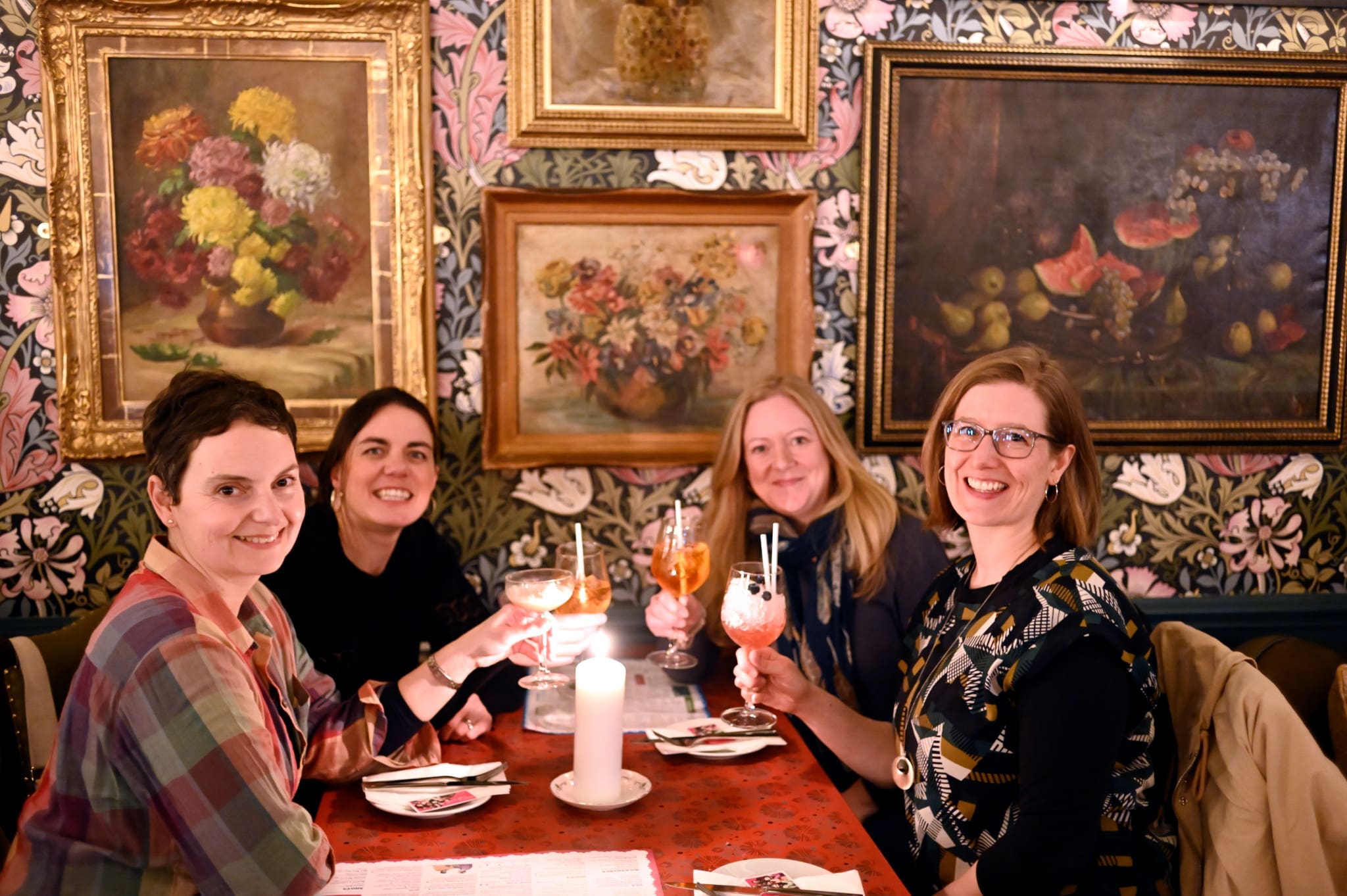Four women sit around a red table in a restaurant, smiling and holding cocktails. The wall behind them features floral wallpaper and framed paintings. A lit candle rests on the table alongside plates of delicious vitello.