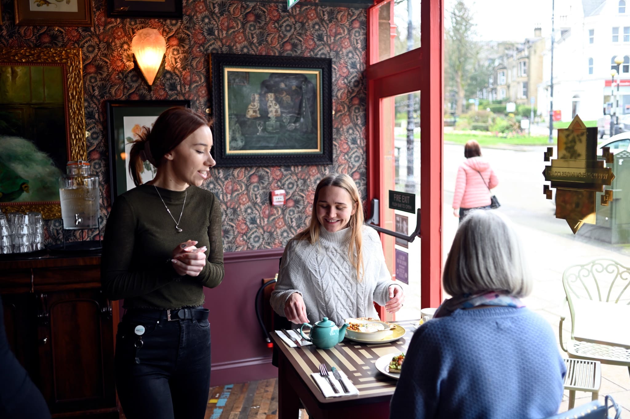 A waitress stands beside a table where two women sit, one smiling with a plate of vitello in front of her. The café has cozy decor, patterned wallpaper, and large windows showing a street scene outside.