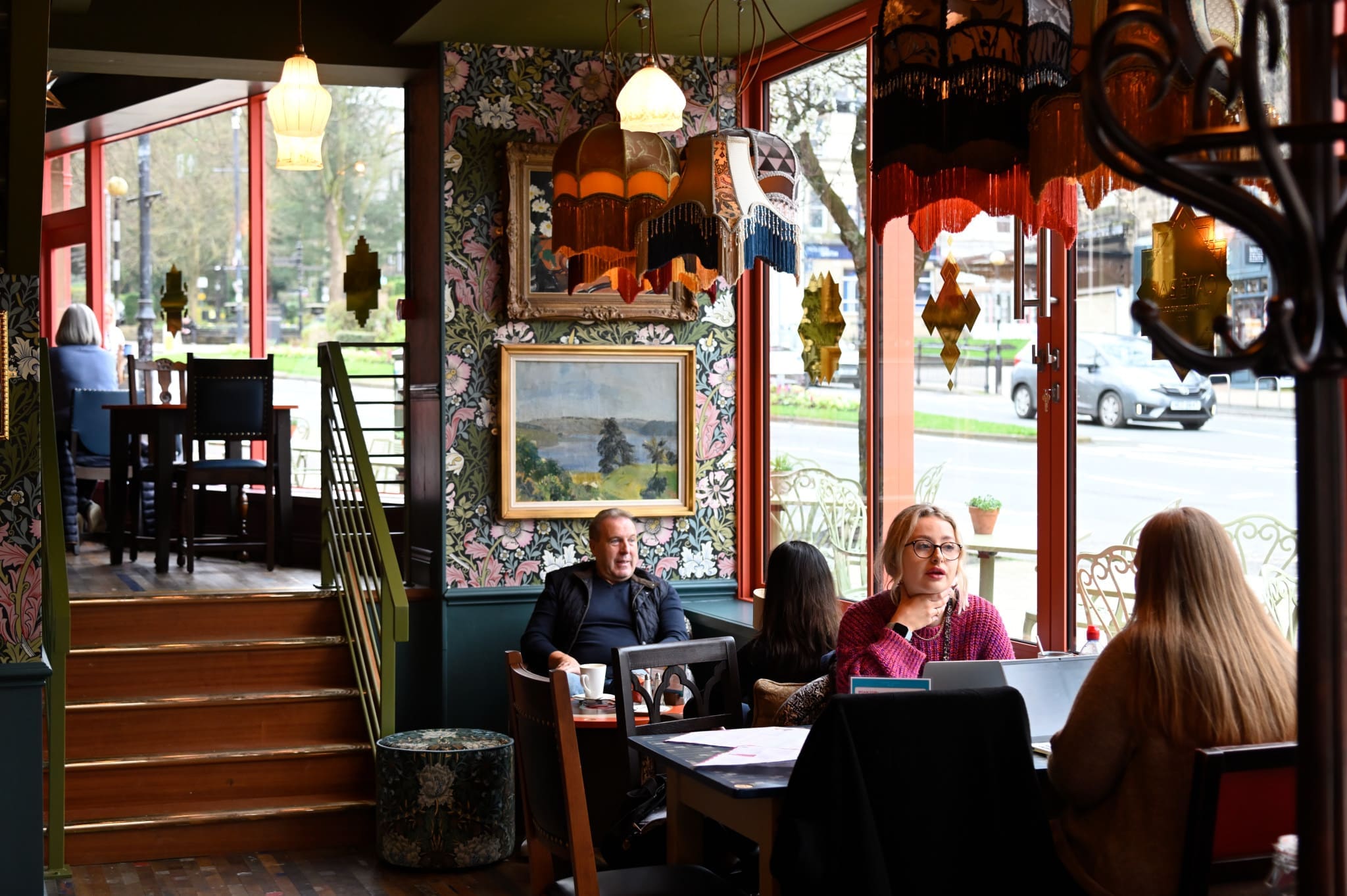 A cozy café with colorful lamps and wallpaper, people chatting and working on laptops near large windows, and a painting hanging on the wall. Natural light fills the space as guests enjoy coffee and delicious vitello dishes.