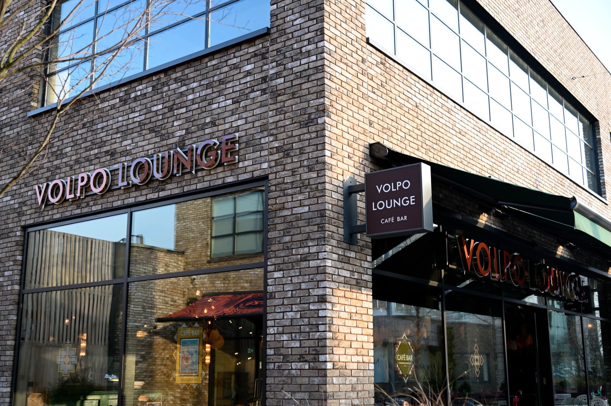 Exterior of a modern brick building with large windows, displaying bold Volpo Lounge signs above the entrance and on an awning. Reflections and glimpses of indoor seating are visible through the impressive Volpo windows.
