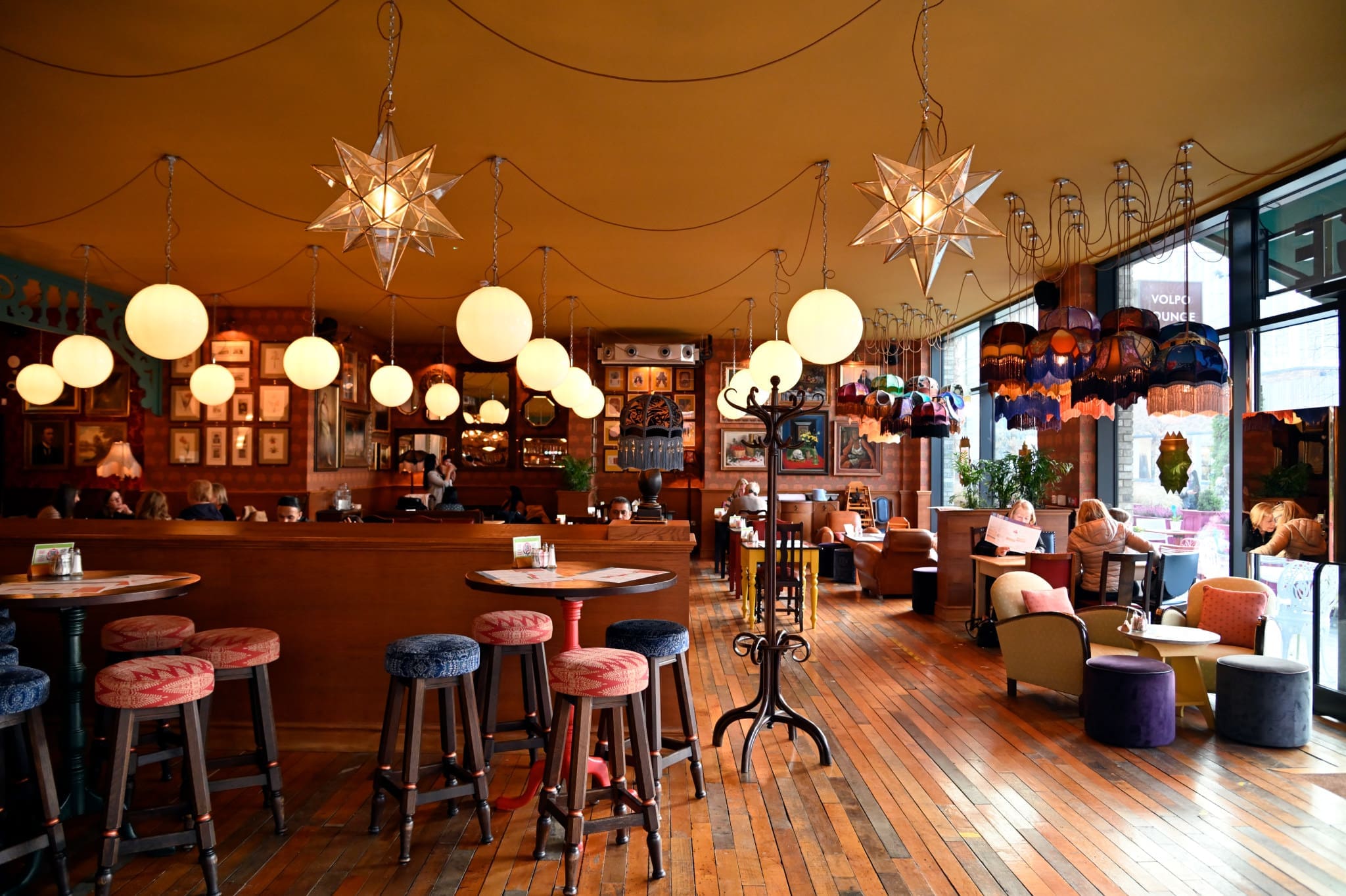 A cozy cafe with wooden floors, round tables and stools, globe and star-shaped hanging lights, eclectic lampshades, large windows, and a few people sitting and chatting by the window at Volpo.