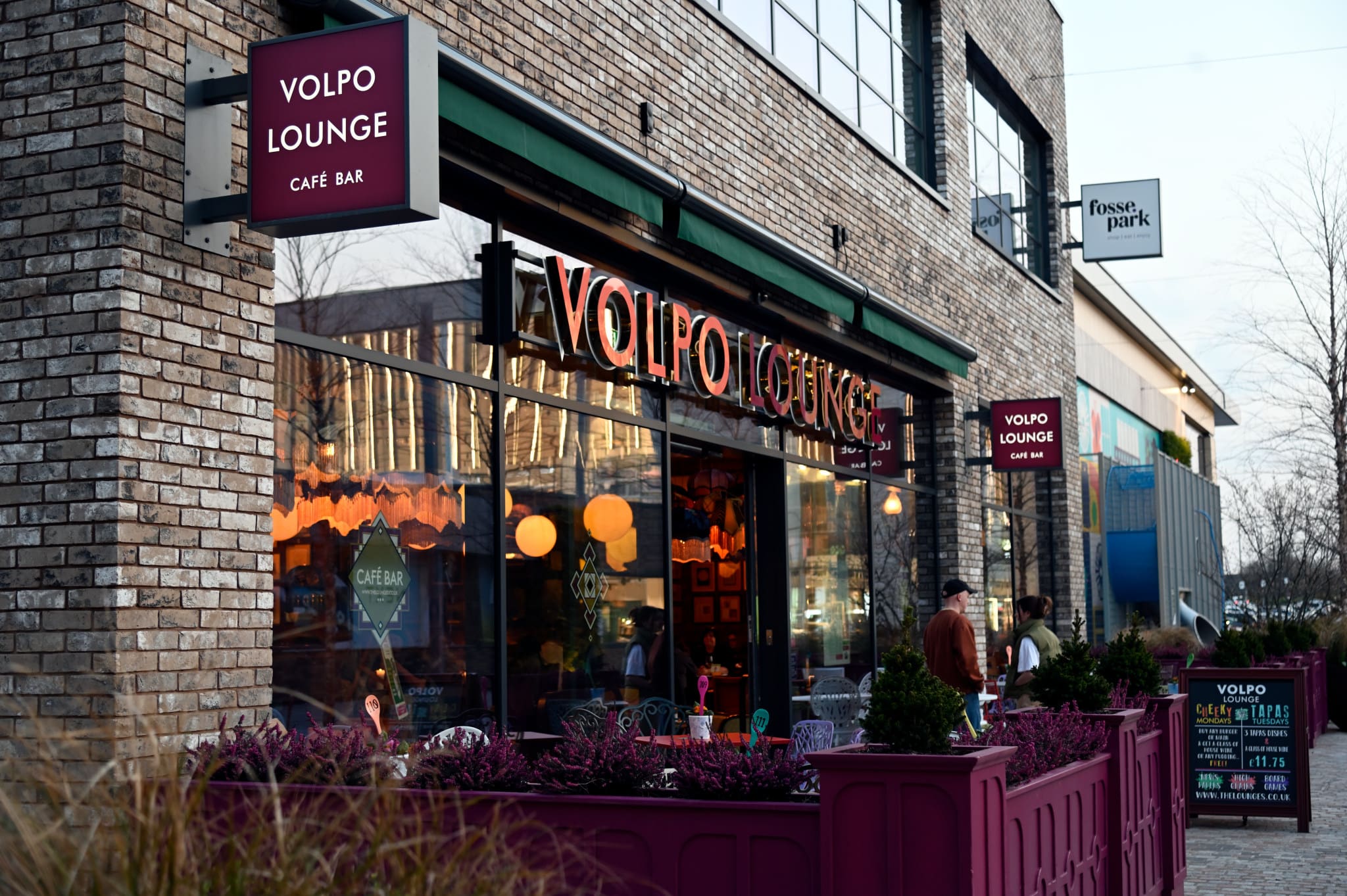 Street view of Volpo Lounge Café Bar with its brick exterior, large windows, and outdoor seating featuring purple planters. People are entering Volpo, and a signboard stands near the entrance.