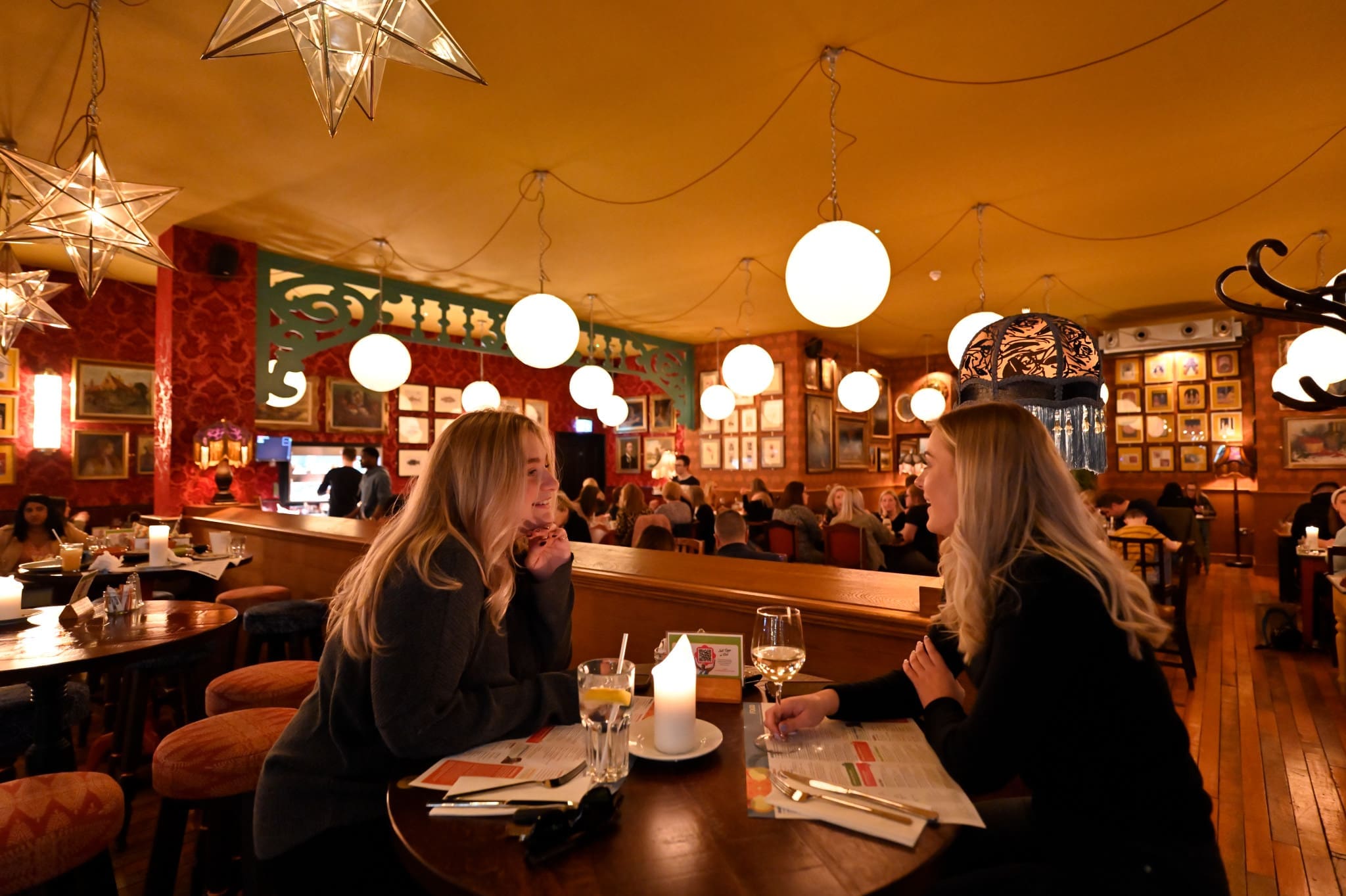 Two women sit at a round table in Volpo, a warmly-lit, cozy restaurant with hanging globe lights and star-shaped lamps, chatting over menus and drinks. Other diners and framed pictures decorate the background.