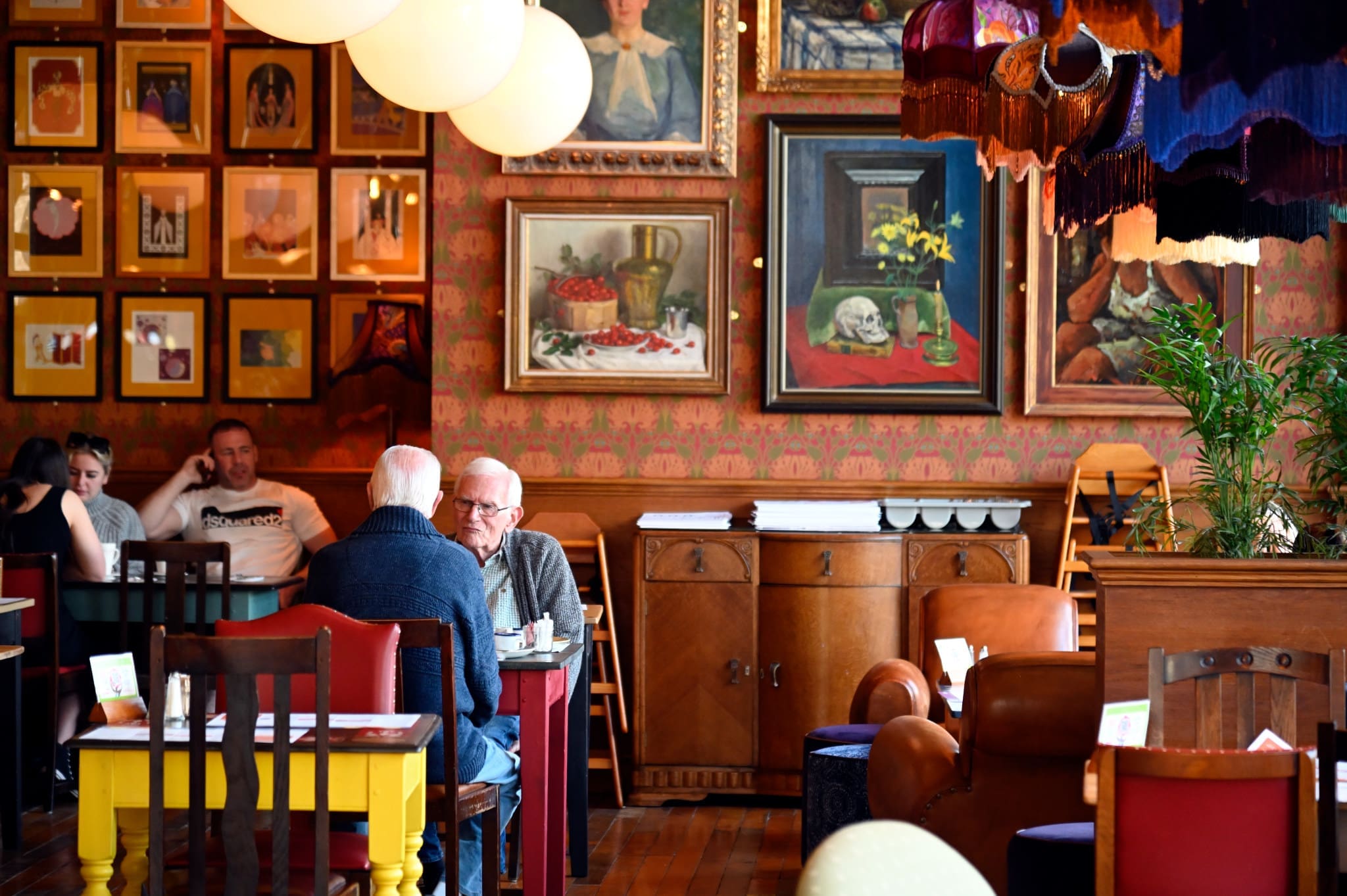 Two older men sit and talk at a small table in Volpo, a cozy, colorful café filled with eclectic framed artwork and vintage decor. Other patrons are visible in the background, and warm lighting creates an inviting atmosphere.