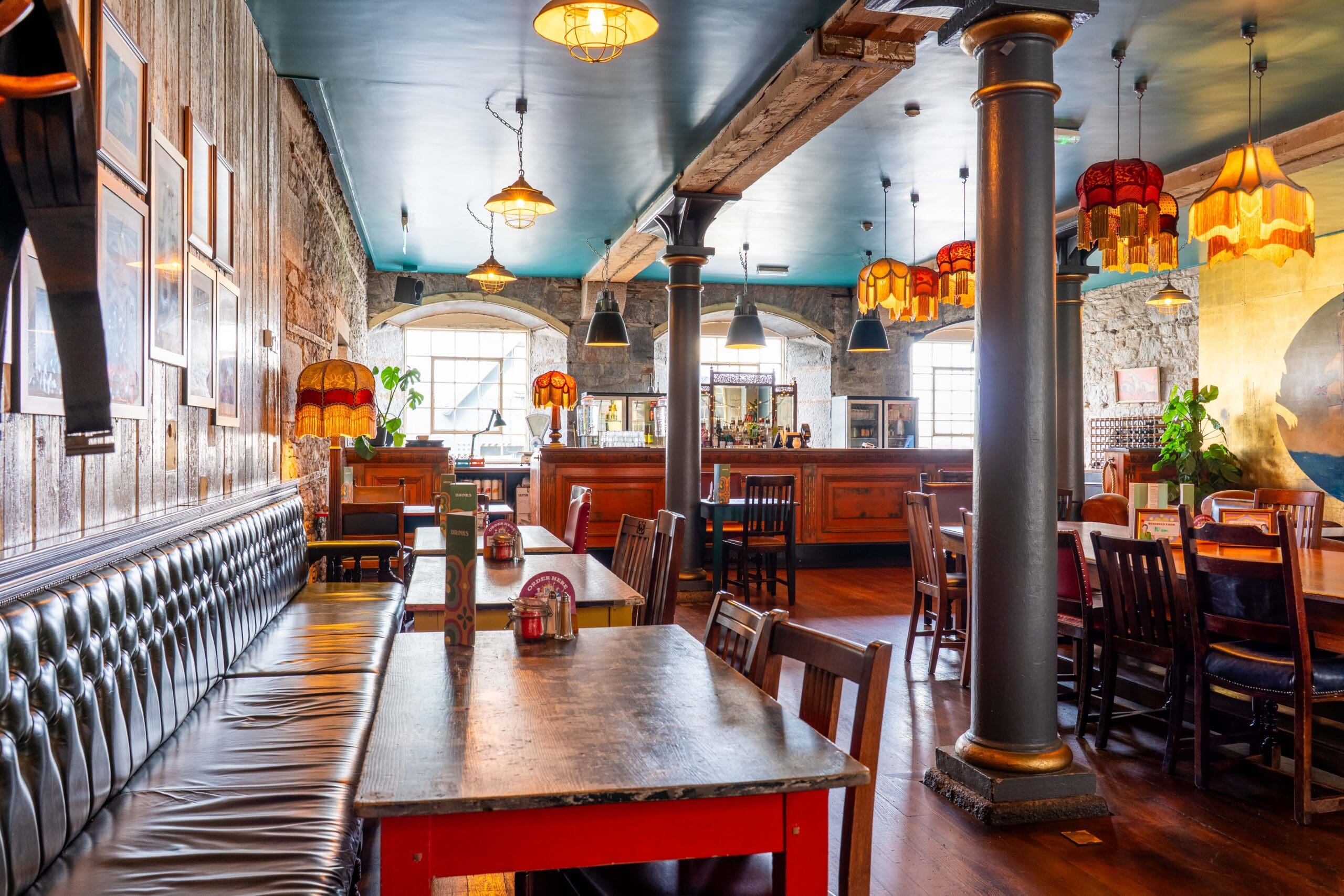 Eclectic restaurant interior with exposed brick walls, vintage lamps, leather banquettes, wooden tables, and chairs. Sunlight streams through large windows, creating a warm, inviting atmosphere.