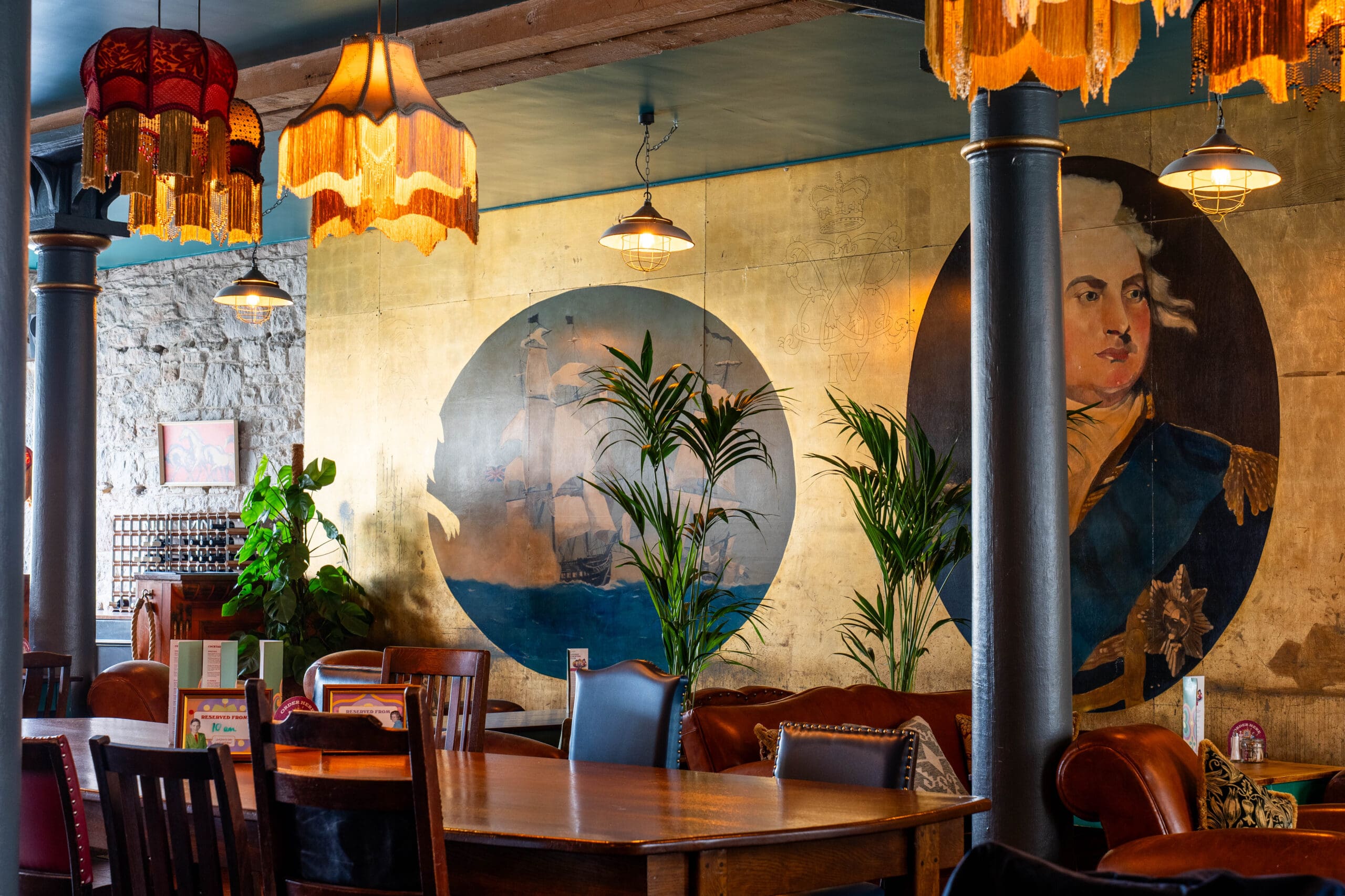 A cozy, eclectic restaurant interior with vintage lamps, wooden tables and chairs, potted plants, and a gold wall featuring large painted portraits, including a historical figure in blue uniform.