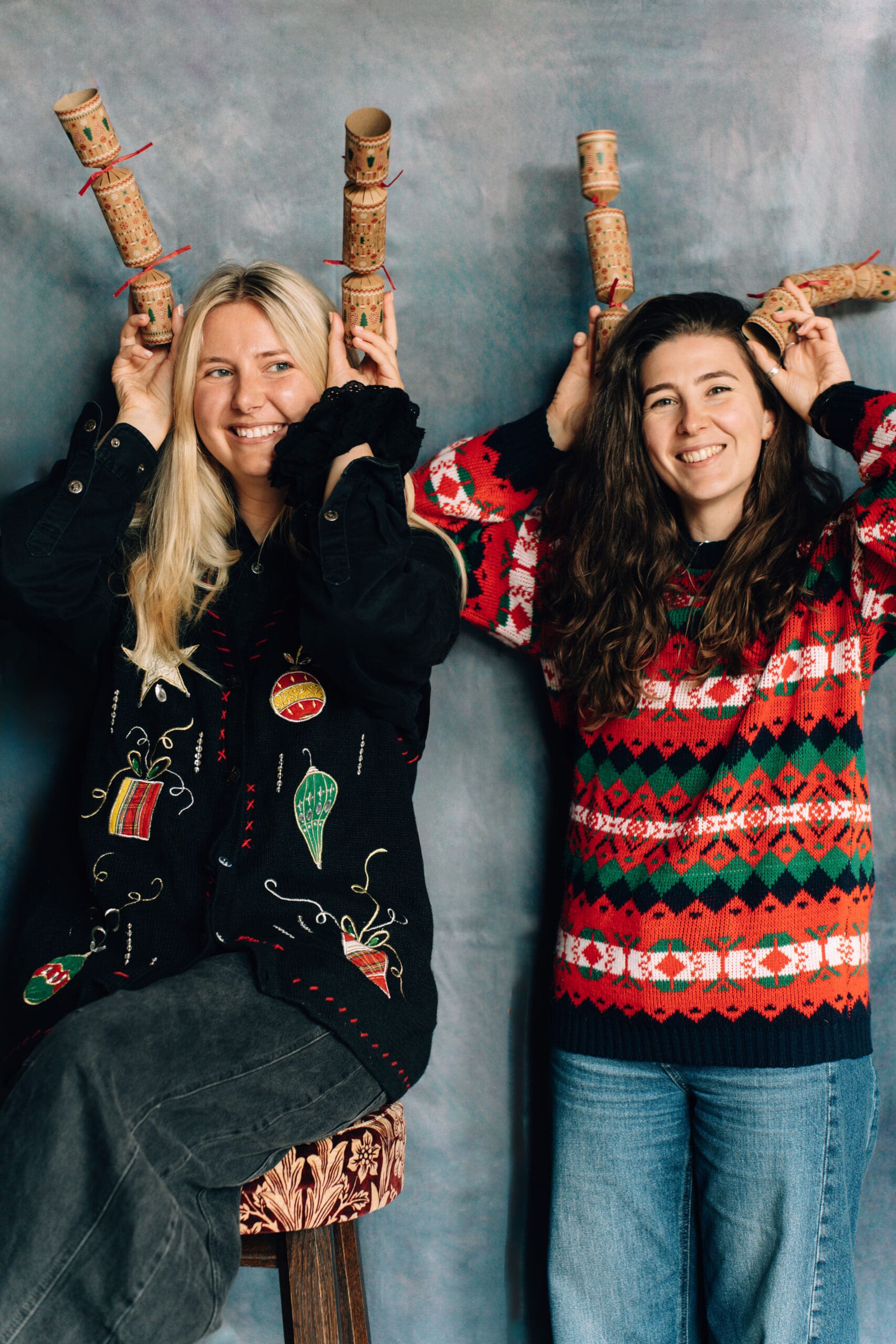 Two women in festive sweaters stand side by side, smiling and holding cork crafts above their heads like reindeer antlers. One sits on a stool while the other stands, both posing playfully for a joyful Christmas photo in front of a neutral backdrop.