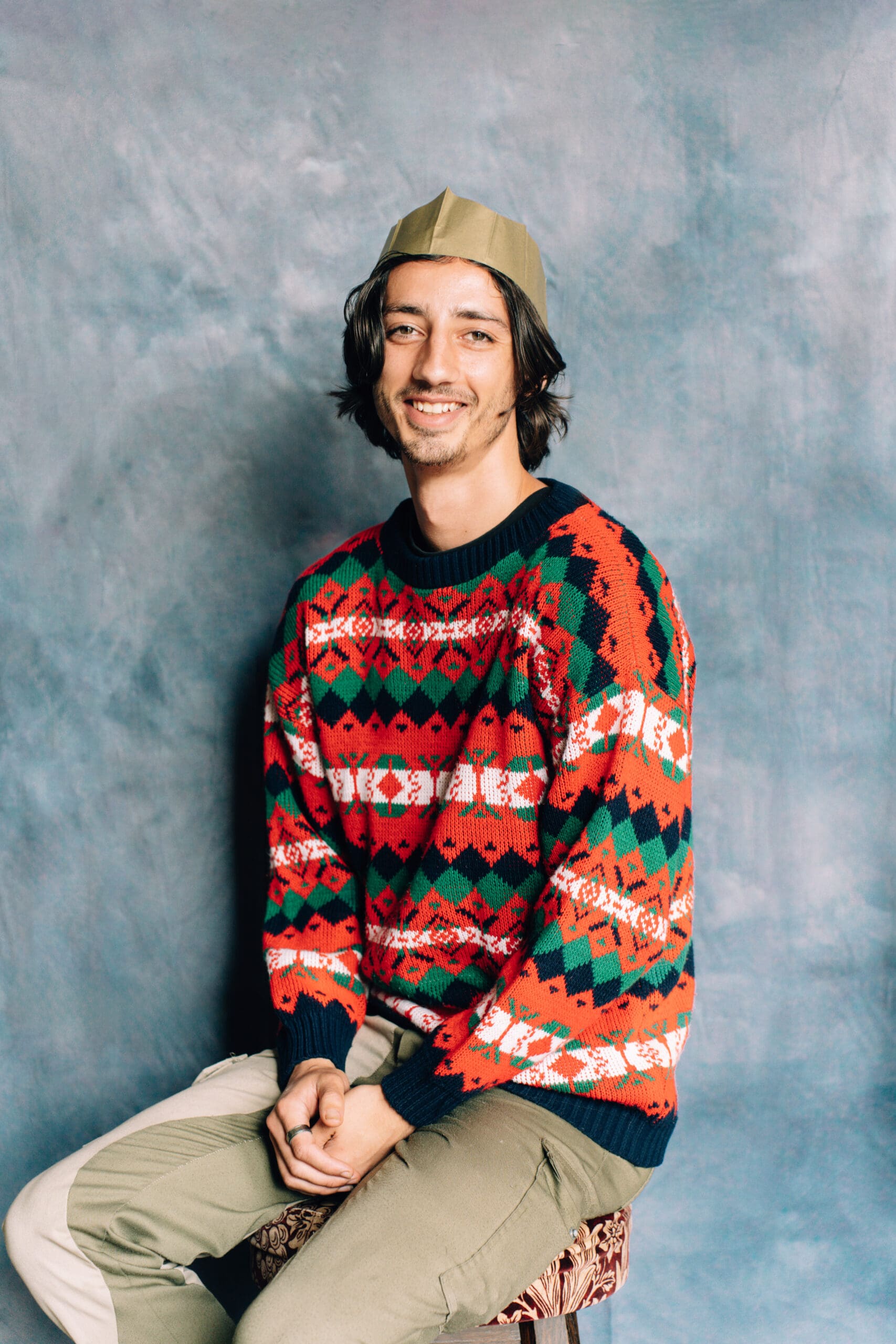 A young man with shoulder-length hair wearing a patterned, colorful knit Christmas sweater, light pants, and a paper crown sits smiling on a stool against a textured blue-gray background.