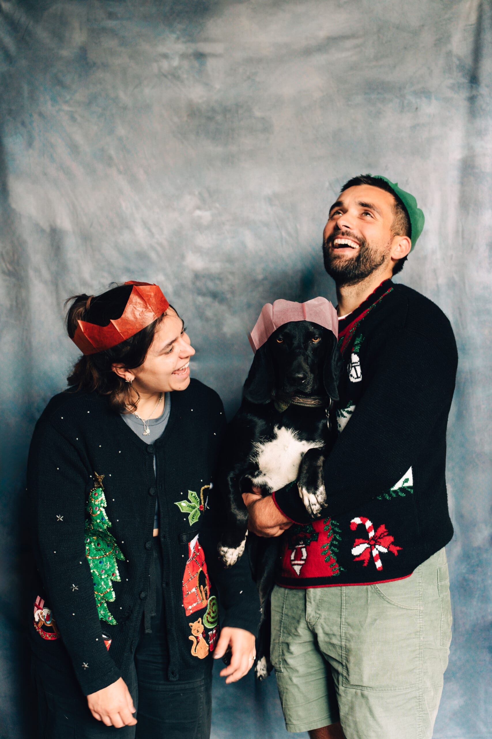 Two people in festive Christmas sweaters and colorful paper crowns smile and laugh while holding a black and white dog, who is also wearing a crown, in front of a gray backdrop.